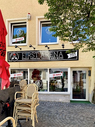 gepflasterte Straße, gelbes Gebäude, schwarzes Schild "First Arena, Kebab, Veggies, Coffee and more", gestapelte Stühle, roter Schirm