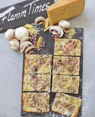 Flammkuchen belegt mit Speck, Pilzen und Käse auf Schieferplatte dekoriert mit ganzen und geschnittenen Pilzen, Speckwürfeln und ganzen und geriebenen Käse; oben links steht auf einer weiteren Schiefernplatte "Flamm Times"
