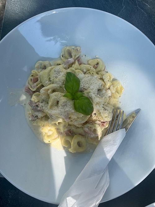 Runder weißer Teller mit Nudeln mit heller Sauce, und Schinkenscheiben, dekoriert mit Parmesan und Basilikum; Besteck eingepackt in einer weißen Serviette