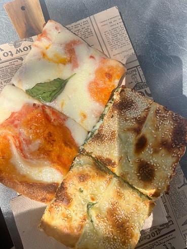 2 Pizzastücke mit Tomaten und viel Käse, 2 Teigstücke mit Spinat-Käse-Füllung überzogen mit Sesam auf einem Schneidebrett aus Holz mit Zeitungspapier und Serviette