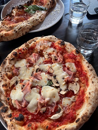 runde Pizza belegt mit Tomatensauce, Käse, Pilzen und Schinken; 2 Gläser Wasser und eine weitere Pizza im Hintergrund
