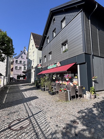 Gepflasterte Straße, dunkles Gebäude mit roter Markise und Stühlen und Tischen