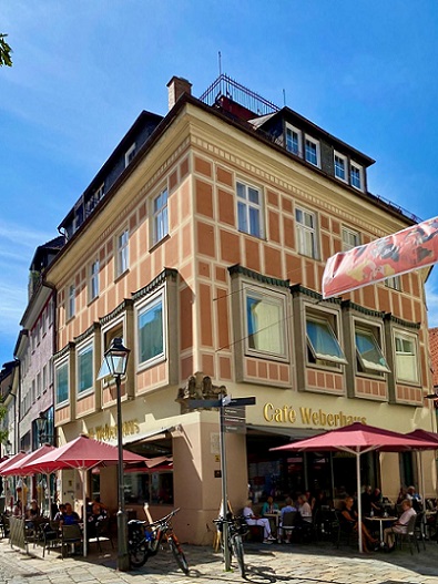 rosa Gebäude, rote Schirme, Personen, Stühle, Laternen, blauer Himmel