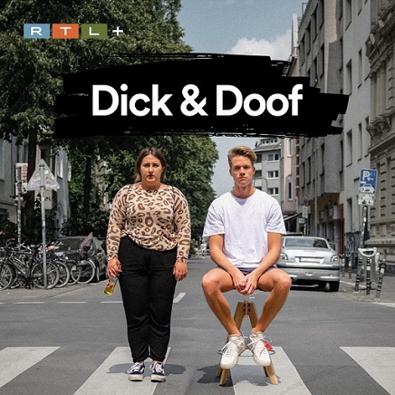 Dick&Doof 2 Personen auf einem Zebrastreifen in einer Stadt. beide sind zur Kamera gerichtet. Die Frau steht und der Mann sitzt auf einem Hocker. Über ihnen steht in weißer Schrift auf schwarzem Hintergrund: "Dick&Doof"
