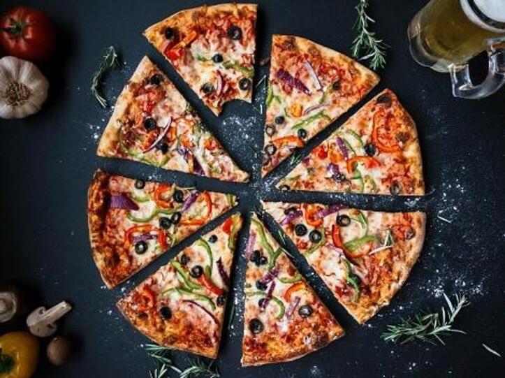 gschnittene Pizza mit einem angebissenen Stück, dunkler Untergrund, Kräuter, Knoblauchknolle, Tomate also Deko außenrum